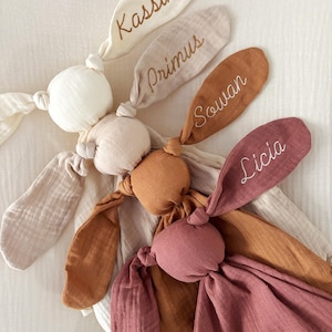 May include: A collection of soft, knotted bunny rabbit plush toys in various neutral colors. Each bunny has long ears with embroidered names: Kassim, Primus, Souan, and Licia. The toys are made of a textured fabric, perfect for a baby.