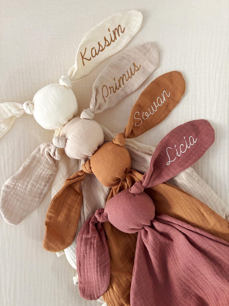 May include: A collection of soft, knotted bunny rabbit plush toys in various neutral colors. Each bunny has long ears with embroidered names: Kassim, Primus, Souan, and Licia. The toys are made of a textured fabric, perfect for a baby.