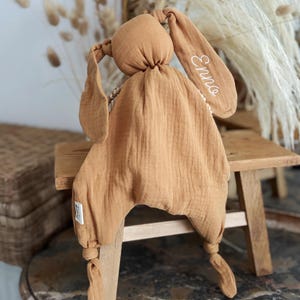 May include: A tan-colored bunny-shaped baby comforter with long floppy ears. The ear on the right has the name "Enno" embroidered in white. The comforter is made of soft, textured fabric and has knotted legs.
