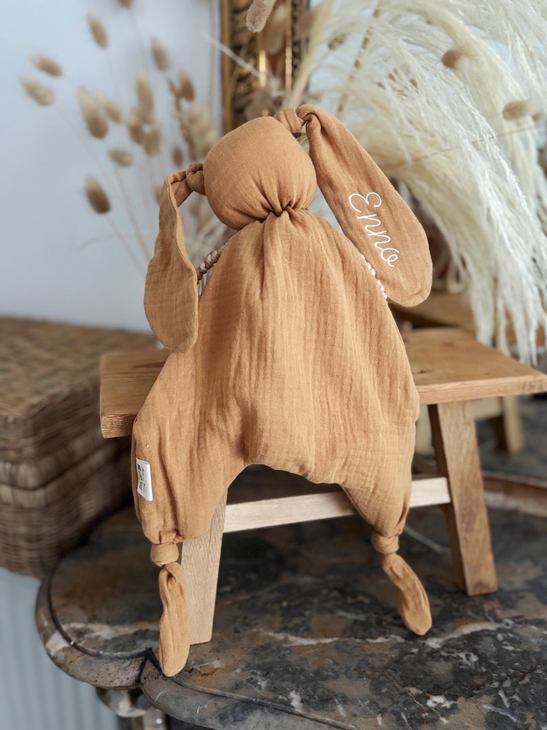 May include: A tan-colored bunny-shaped baby comforter with long floppy ears. The ear on the right has the name "Enno" embroidered in white. The comforter is made of soft, textured fabric and has knotted legs.