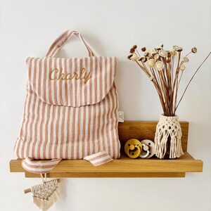 May include: A striped peach and white fabric backpack with the name Charly embroidered in gold thread on the flap. A small macrame charm hangs from the strap. Beside it are two round pacifiers and a vase of dried flowers in a macrame holder.