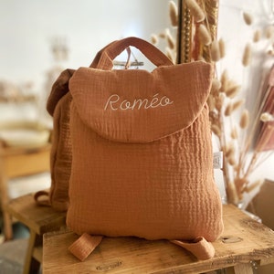 May include: A rust-coloured cotton muslin backpack featuring a top handle and a flap closure. The name Rom&eacute;o is embroidered in white cursive script on the front flap. The backpack has a textured, crinkled fabric finish and two shoulder straps.
