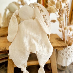 May include: Cream-colored bunny-shaped baby comforter. The comforter has long, floppy ears with the embroidered text "petit beignet" in a gold color. The body is a soft, quilted fabric with knotted feet. The comforter is on a wooden surface.