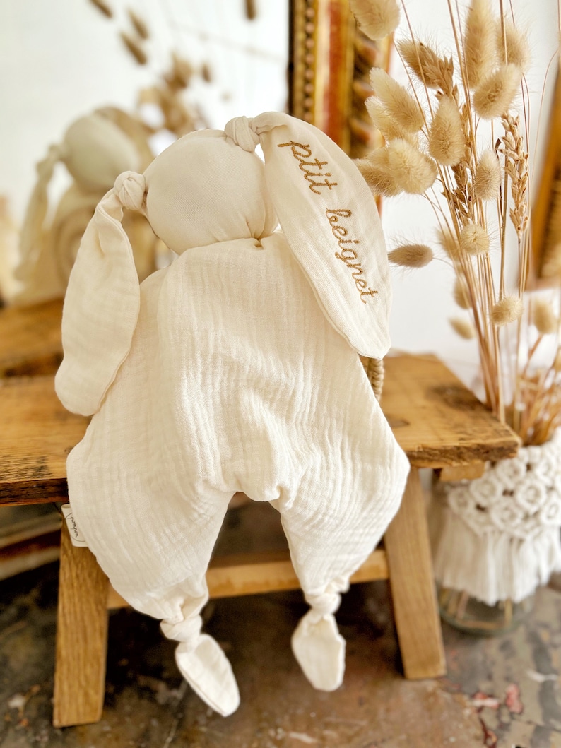 May include: Cream-colored bunny-shaped baby comforter. The comforter has long, floppy ears with the embroidered text "petit beignet" in a gold color. The body is a soft, quilted fabric with knotted feet. The comforter is on a wooden surface.