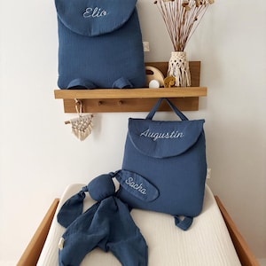 May include: Two blue fabric backpacks with top handles and flap closures, each embroidered with names Elio and Augustin. A matching blue fabric bunny-shaped security blanket with the name Sacha embroidered on one ear is included.