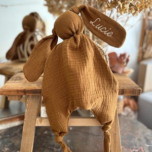 May include: A mustard-colored bunny-shaped baby comforter with long floppy ears. The right ear has the name "Lucie" embroidered in white. The comforter is made of a soft, textured fabric and is knotted at the bottom.