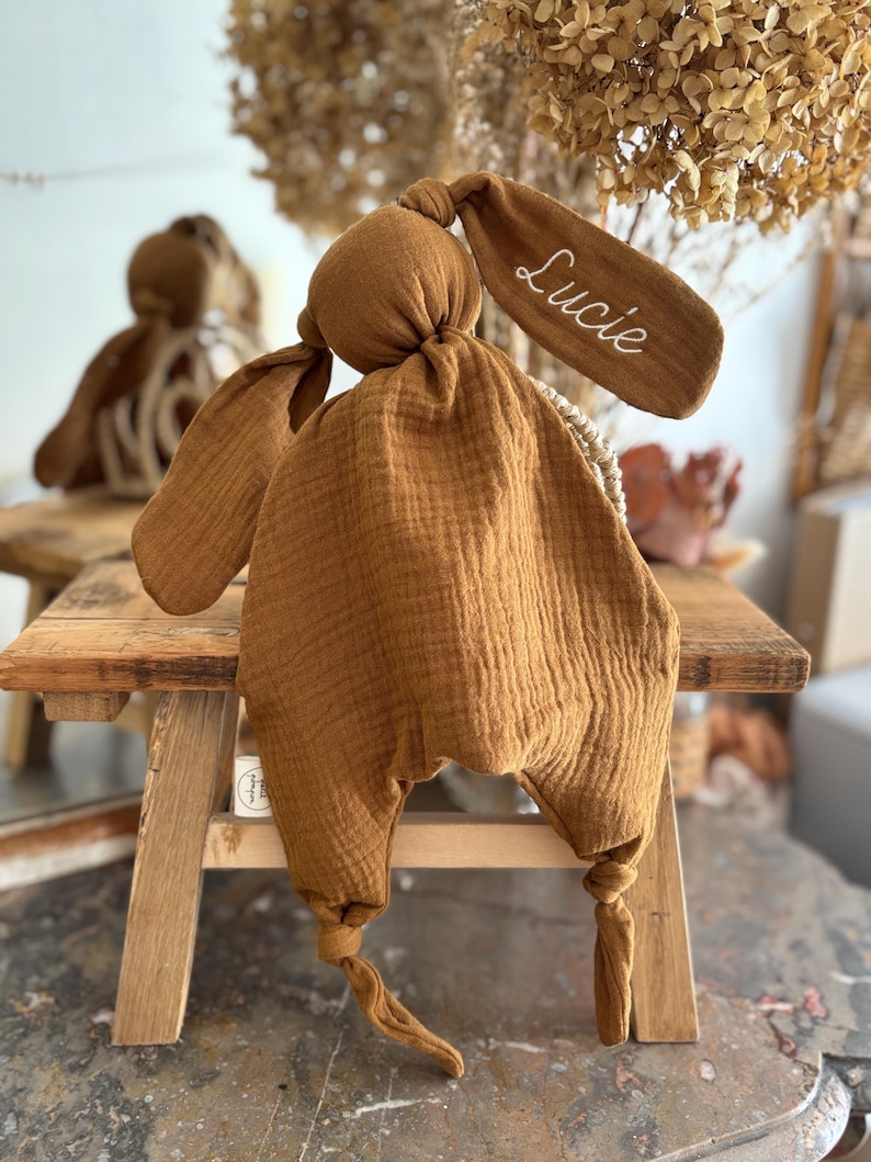 May include: A mustard-colored bunny-shaped baby comforter with long floppy ears. The right ear has the name "Lucie" embroidered in white. The comforter is made of a soft, textured fabric and is knotted at the bottom.