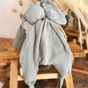 May include: A soft, gray bunny-shaped baby comforter with long, floppy ears. The name "Romy" is embroidered in white on one ear. The comforter is made of a textured fabric and has knotted feet.