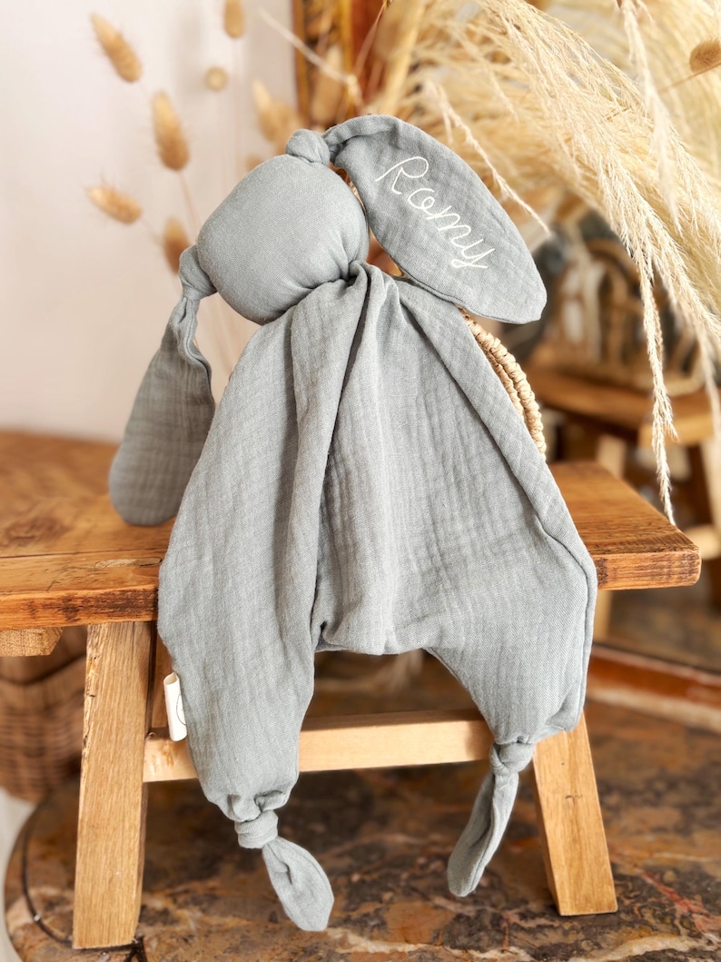 May include: A soft, gray bunny-shaped baby comforter with long, floppy ears. The name "Romy" is embroidered in white on one ear. The comforter is made of a textured fabric and has knotted feet.