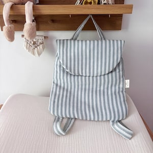 May include: A soft, padded backpack featuring a vertical striped pattern in light blue and white. The bag has a rounded top flap, a single top handle, and two adjustable shoulder straps. A small white rectangular brand tag is visible on the side.