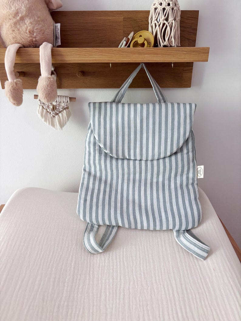May include: A soft, padded backpack featuring a vertical striped pattern in light blue and white. The bag has a rounded top flap, a single top handle, and two adjustable shoulder straps. A small white rectangular brand tag is visible on the side.