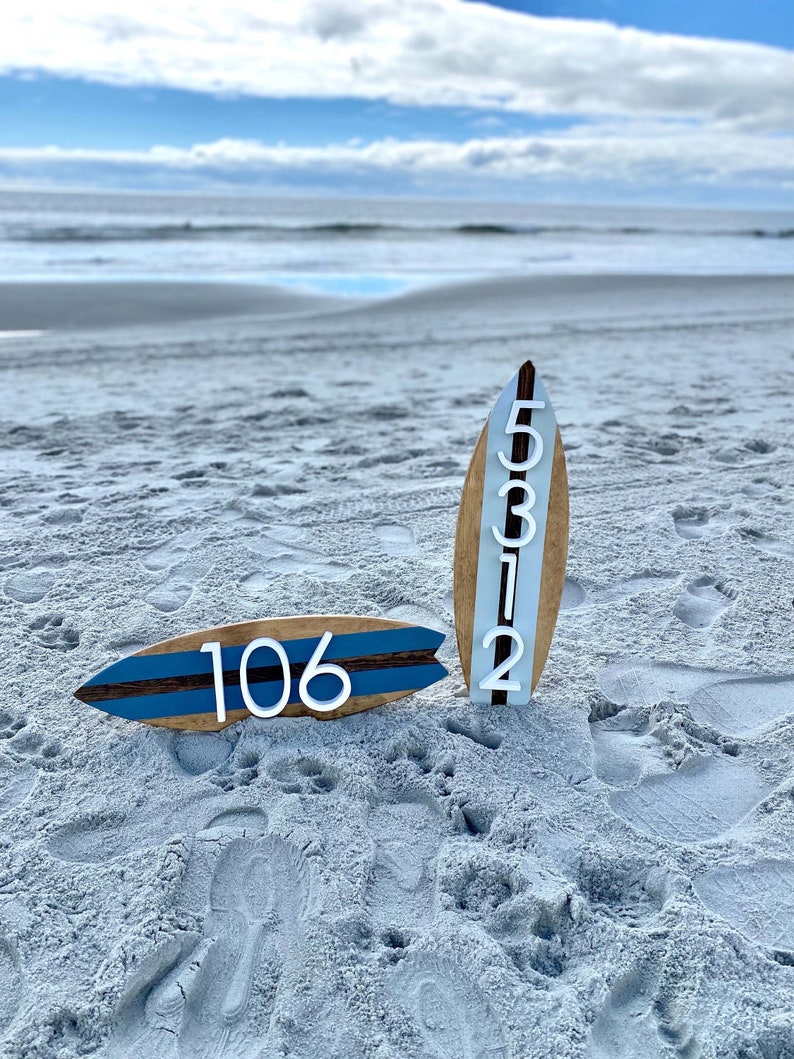 Puede incluir: Dos tablas de surf de madera con n&uacute;meros blancos en ellas. La tabla de surf de la izquierda tiene el n&uacute;mero 106. La tabla de surf de la derecha tiene los n&uacute;meros 5312. Las tablas de surf est&aacute;n en una playa de arena.