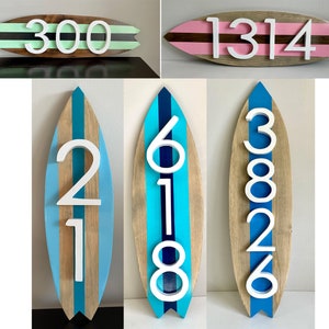 Puede incluir: Cuatro letreros de n&uacute;meros de casa de madera con forma de tabla de surf. El primer letrero es verde con rayas negras y el n&uacute;mero 300. El segundo letrero es rosa con rayas marrones y el n&uacute;mero 1314. El tercer letrero es marr&oacute;n con rayas azules y el n&uacute;mero 21. El cuarto letrero es marr&oacute;n con rayas azules y el n&uacute;mero 618.