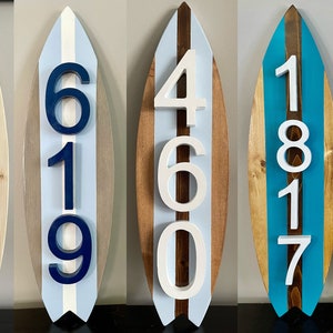 Puede incluir: Cinco letreros de n&uacute;meros de casa de madera con forma de tabla de surf con pintura blanca, azul y marr&oacute;n. Los n&uacute;meros son 12740, 619, 460, 1817 y 939.