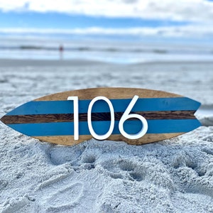 Puede incluir: Una tabla de surf de madera con rayas azules y marrones pintadas en ella. El n&uacute;mero 106 est&aacute; escrito en blanco en la tabla de surf. La tabla de surf est&aacute; sentada en una playa de arena.