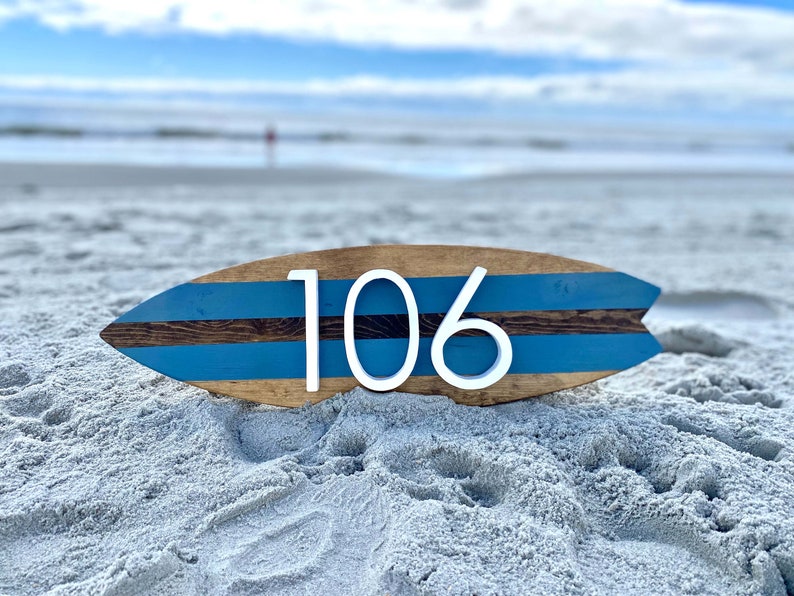 Puede incluir: Una tabla de surf de madera con rayas azules y marrones pintadas en ella. El n&uacute;mero 106 est&aacute; escrito en blanco en la tabla de surf. La tabla de surf est&aacute; sentada en una playa de arena.