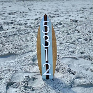 Puede incluir: Una tabla de surf de madera con una franja azul claro en el centro. Los n&uacute;meros 5312 est&aacute;n pintados en blanco en la tabla de surf.