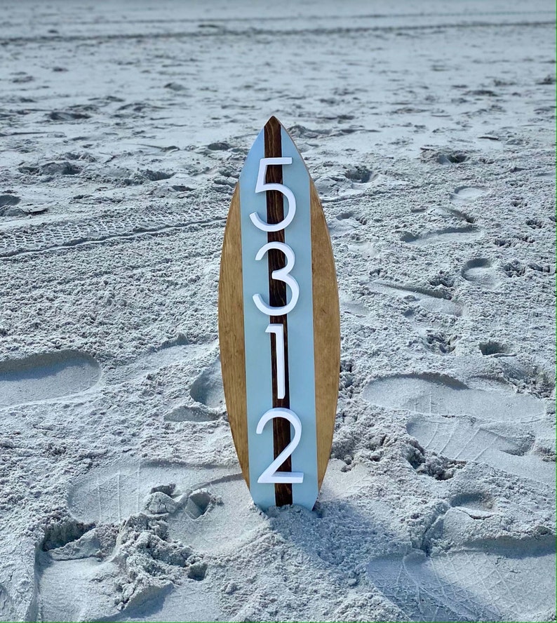 Puede incluir: Una tabla de surf de madera con una franja azul claro en el centro. Los n&uacute;meros 5312 est&aacute;n pintados en blanco en la tabla de surf.