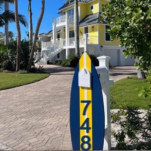 May include: A surfboard-shaped mailbox post featuring a vertical yellow stripe centered between two dark blue panels. The number 748 is displayed in large blue digits on the lower section, with a white mailbox mounted at the top of the post.
