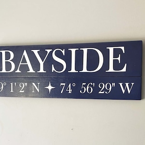 May include: A navy blue wooden sign with white lettering that reads "BAYSIDE" and the coordinates "39° 1' 2" N 74° 56' 29" W".