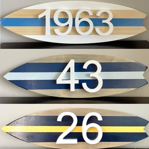 Puede incluir: Tres letreros de n&uacute;meros de casa de madera con forma de tabla de surf con rayas azules, blancas y amarillas. Los n&uacute;meros son 1963, 43 y 26.