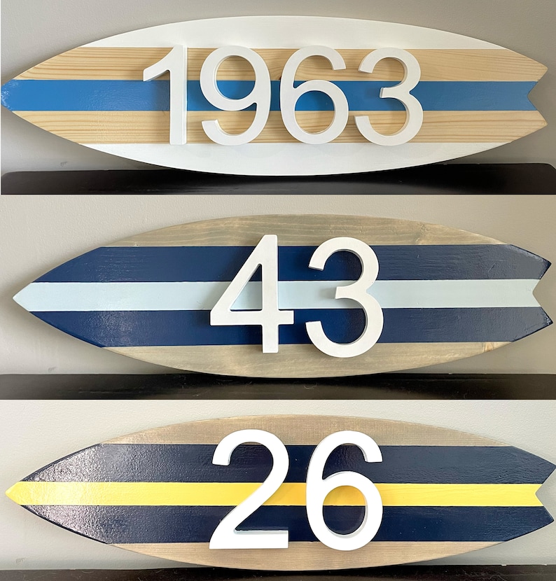 Puede incluir: Tres letreros de n&uacute;meros de casa de madera con forma de tabla de surf con rayas azules, blancas y amarillas. Los n&uacute;meros son 1963, 43 y 26.
