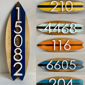 Puede incluir: Cinco letreros de n&uacute;meros de casa de madera con forma de tabla de surf con rayas de colores diferentes y n&uacute;meros blancos. Los n&uacute;meros son 210, 4468, 116, 6605 y 204.