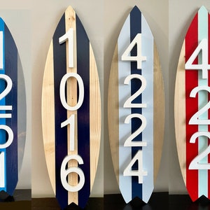 Puede incluir: Tres letreros de n&uacute;meros de casa de madera con forma de tabla de surf con n&uacute;meros blancos. El primer letrero es azul y gris con los n&uacute;meros 1251. El segundo letrero es de madera con rayas azules y blancas y los n&uacute;meros 1016. El tercer letrero es rojo y blanco con los n&uacute;meros 4224.