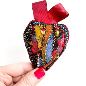 Peut inclure: Un petit coussin en forme de cœur avec un motif patchwork dans des tons de rouge, bleu, jaune et noir. Le coussin est doté d'un ruban rouge attaché au sommet.