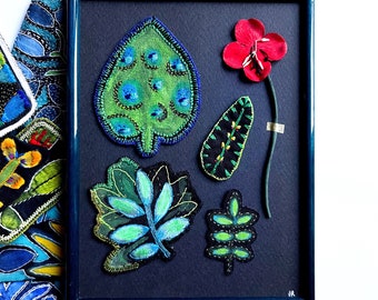 以下が含まれることがあります： 刺繍された布の葉と赤い花が特徴の額装されたアートワーク。葉はさまざまな緑と青の色合いで、複雑なステッチの詳細があります。フレームは濃い青色で、背景も同じ濃い青色です。