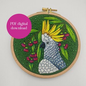 Könnte beinhalten: Gesticktes Bild in einem Holzrahmen, das einen weißen und blauen Kakadu mit gelbem Kamm, grünen Blättern und rosa Blüten zeigt. Der Hintergrund ist grüner Stoff. Der Text "PDF digital download" befindet sich auf einem rosa Kreis.