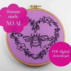 May include: A circular wooden embroidery hoop containing purple fabric with a black thread outline of a bumblebee centered within a heart-shaped floral wreath. Pink circular text overlays read Human made NO AI and PDF digital download.