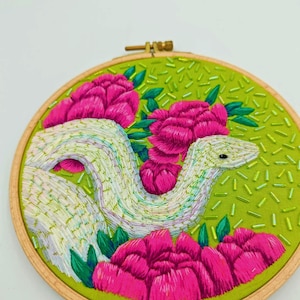May include: Embroidered artwork featuring a white snake with a textured pattern, surrounded by vibrant pink flowers and green leaves. The background is a light green fabric with small green beads, all set within a wooden embroidery hoop.