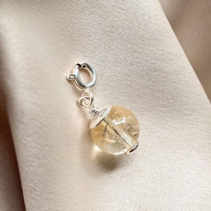 May include: A silver charm with a clear, light yellow, round gemstone. The charm has a silver clasp.