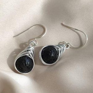 Puede incluir: Un par de pendientes envueltos en alambre de plata. Cada pendiente presenta una piedra negra esférica, envuelta en alambre de plata enrollado, con un gancho simple para la oreja. Los pendientes se muestran sobre una superficie satinada de color beige claro.