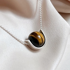 Peut inclure: Un collier en argent avec une pierre précieuse œil de tigre polie et ronde. La pierre est sertie dans un support argenté incurvé et suspendue à une fine chaîne en argent. L'œil de tigre présente des bandes marron et dorées.