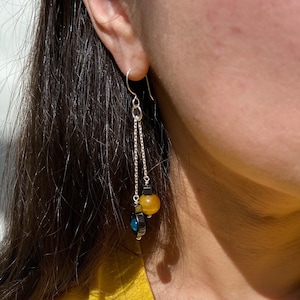 Op de afbeelding: Hangende oorbellen met zilverkleurige kettingen en haken. Elke oorbel heeft een gele kraal, een donkerblauwe en zilverkleurige kraal en een kleinere gele kraal. De oorbellen worden getoond tegen een gele top.