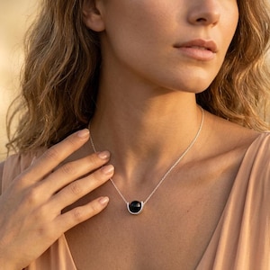 Pode incluir: Um colar de prata com uma única pedra preta redonda e lisa, inserida num suporte de prata em forma de U. O pendente está suspenso por uma corrente de prata fina e delicada que repousa sobre a clavícula, criando um visual simples.