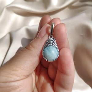 Puede incluir: Un colgante de plata con una piedra preciosa redonda de color azul claro. La piedra está encerrada en una jaula de plata en forma de lágrima. El colgante tiene un pequeño lazo en la parte superior para una cadena. El fondo es de tela blanquecina.