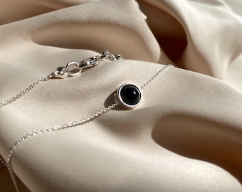 Peut inclure: Un délicat collier en argent avec un petit pendentif rond en pierre noire. Le pendentif est serti dans une lunette en argent et est suspendu à une fine chaîne en argent. Le collier est présenté sur un tissu satiné crème.