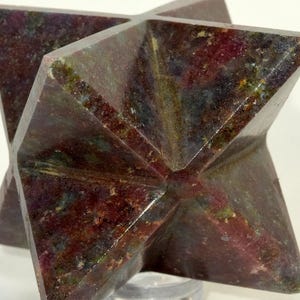 May include: A multi-faceted, star-shaped sculpture. The stone is a deep burgundy color with flecks of green and gray. The geometric design is displayed on a clear, circular stand.