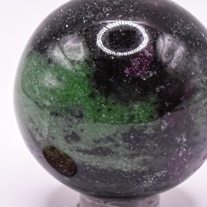 May include: A polished spherical ruby in zoisite gemstone featuring deep green, black, and magenta-pink mineral inclusions. The sphere is positioned next to a copper-colored one-cent coin for scale, showing the relative size of the stone.