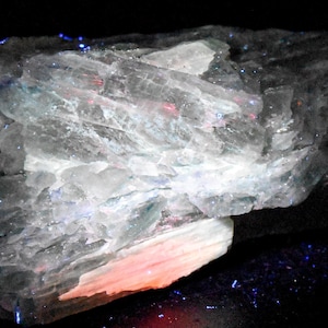 May include: A mineral specimen under UV light, displaying a range of colors. The rock formation has a crystalline structure with areas of blue, red, and white. The text at the top reads "Under LW UV Light 365nm w/ Optical UV Filter".