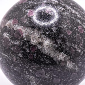 May include: A polished sphere of dark gray stone with flecks of red and white. The spherical object has a smooth surface and is likely a decorative item or a gemstone. The stone's color is a mix of black, gray, and red.
