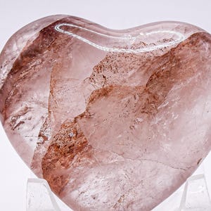 Op de afbeelding: Een hartvormige kristal met een doorschijnende, roze tint en roodbruine insluitsels. De gepolijste steen staat op een heldere standaard, die de natuurlijke patronen benadrukt. Ideaal voor decoratie.