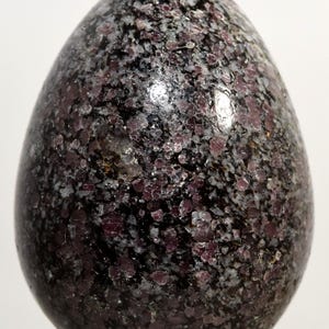 Può includere: Un oggetto decorativo lucido a forma di uovo. La superficie è un mosaico di frammenti minerali grigio scuro, neri e rosso bordeaux intenso. L'uovo ha una finitura liscia e lucida, che riflette la luce. L'oggetto è probabilmente una pietra decorativa o un campione minerale.