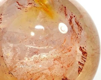 May include: A large, clear quartz sphere with inclusions of red and orange iron oxide. The sphere is polished and has a smooth, glassy surface.