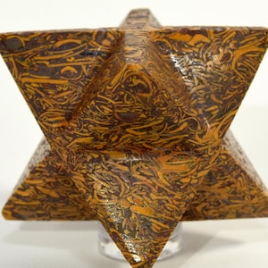 May include: A six-pointed star-shaped sculpture, known as a merkaba, crafted from a polished stone with a complex, swirling pattern of dark brown, tan, and ochre colors. The geometric object features sharp, symmetrical points and smooth surfaces.