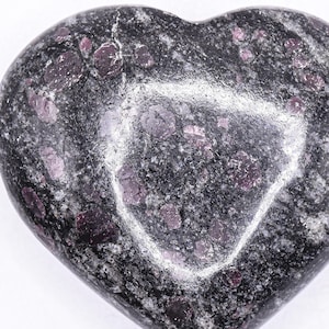 Op de afbeelding: Een hartvormige steen met een gepolijst oppervlak. De steen is voornamelijk donkergrijs met vlekken van lichtgrijs en dieprode insluitsels. Het hart is glad en symmetrisch, met een lichte glans.