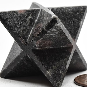 Può includere: Una scultura in pietra a forma di stella, di colore grigio scuro, con una superficie strutturata. L'opera d'arte geometrica è esposta su sfondo bianco accanto a una moneta da un centesimo per il confronto delle dimensioni. La scultura ha un design unico a più punte.